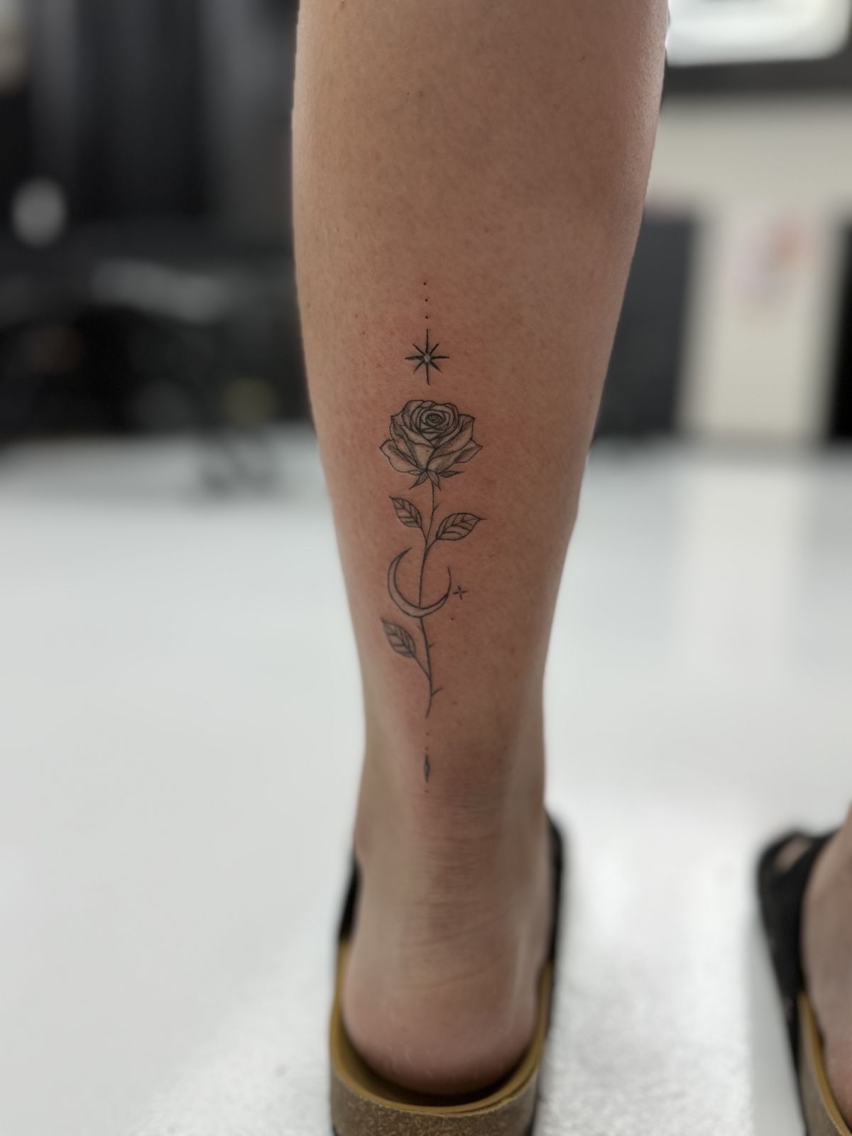 Fine-line rose tattoo with star and crescent moon on the calf, black and grey shading